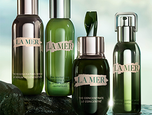 Kategoriebild Seren von La Mer: Verschiedene grüne Glasflakons mit Pump- und Pipettenverschluss stehen auf dunklem Untergrund vor aquatisch beleuchtetem Hintergrund.