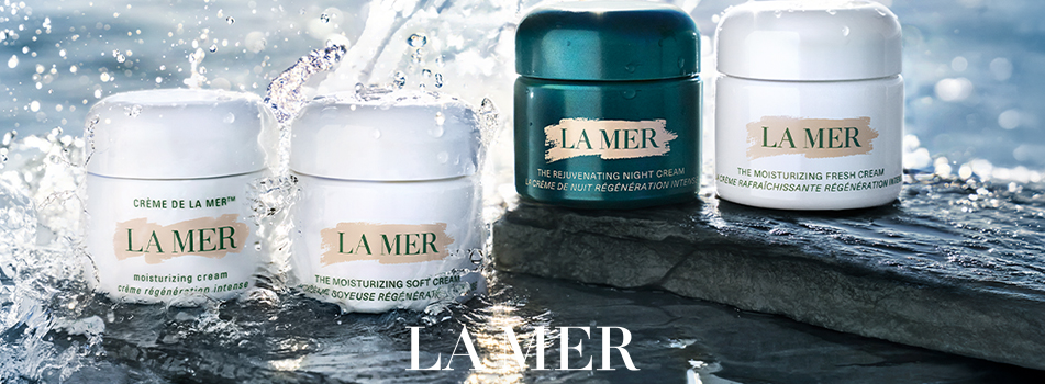 Markenbanner von La Mer: Mehrere Cremetiegel und Pflegeprodukte der Marke La Mer stehen auf nassen Felsen, umgeben von spritzendem Wasser vor maritimem Hintergrund.
