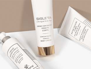 Drei Pflegeprodukte von Sisley Paris auf heller Fläche vor beigem Hintergrund: Im Mittelpunkt eine weiße Tube mit goldfarbenem Deckel und der Aufschrift 'Sisleÿa L'Intégral Anti-Âge – Concentrated Firming Body Cream'. Links und rechts daneben zwei weitere Tuben mit schwarzer Beschriftung und silbernen bzw. weißen Verschlüssen, teilweise angeschnitten.
