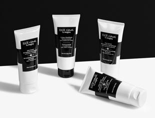 Vier Tuben Haarpflegeprodukte der Linie 'Hair Rituel by Sisley' stehen und liegen auf einer kontrastreichen schwarz-weißen Fläche. Die Tuben sind weiß mit schwarzen Etiketten und enthalten verschiedene Produkte für Haar- und Kopfhautpflege. Die Anordnung wirkt modern und stilisiert vor einem minimalistischen Hintergrund.